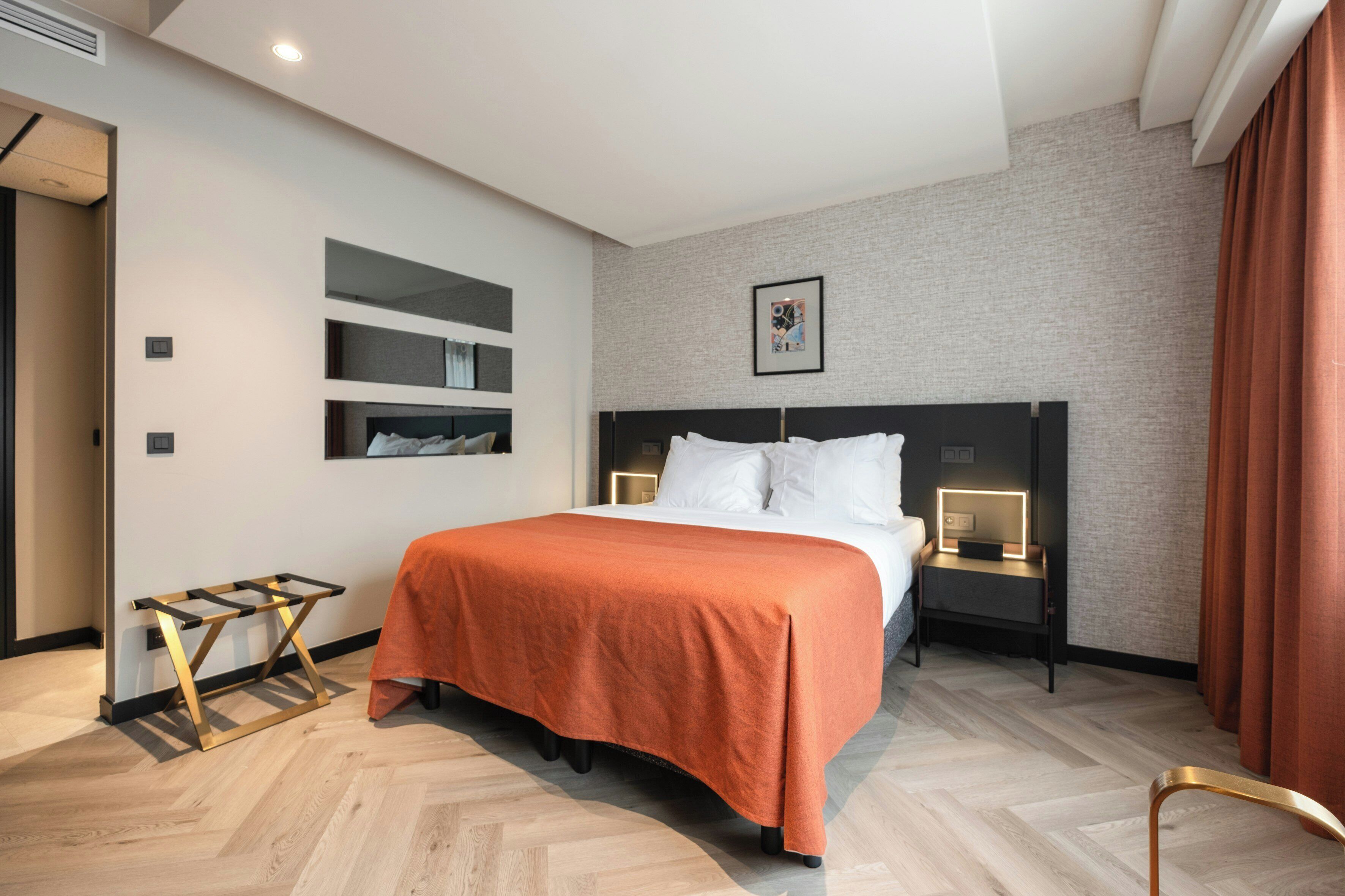 Una habitación de hotel moderna con una cama, una mesita de noche con lámpara, un pequeño estante y suelo de madera.