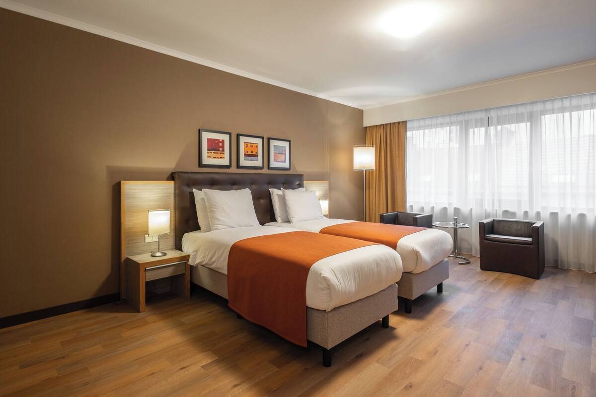 A hotel room with two beds, a bedside table with a lamp, a chair, a small table, and a large window with curtains.
