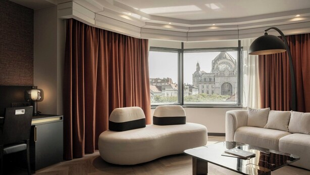 A modern living room with a curved sofa, a glass coffee table, and a view of a historic building through large windows. - Hyllit Hotel (Antwerp)