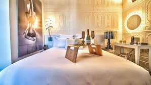 A hotel room with a large bed, a wooden headboard, a wall-mounted mirror, a bedside table with a lamp, and a decorative wall panel.