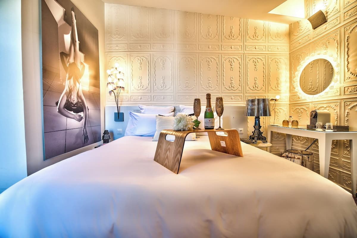 Habitación de hotel con una cama grande, cabecera de madera, espejo montado en la pared, mesita de noche con lámpara y un panel decorativo en la pared.