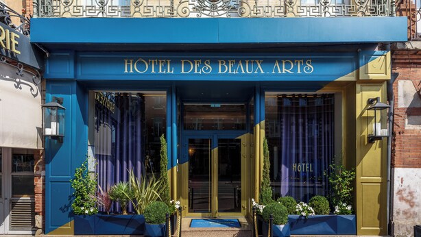 The exterior of HOTEL DES BEAUX ARTS with a blue facade, large glass windows, and a prominent entrance.
