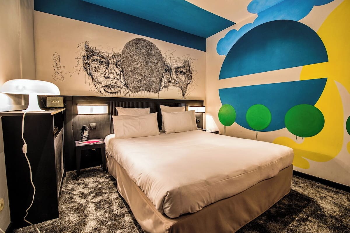 Habitación de hotel con cama, escritorio, lámpara y un muro con un mural colorido.