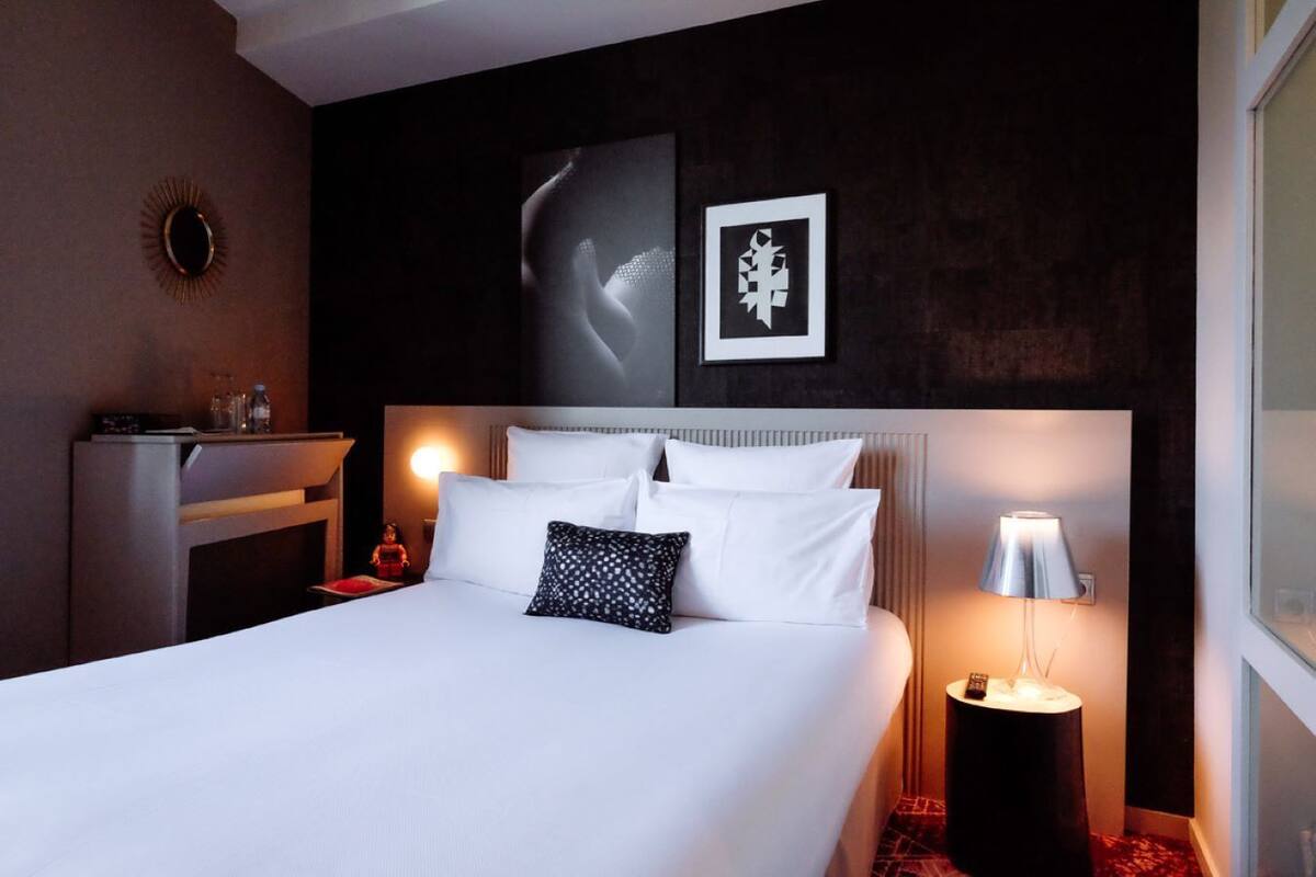 Habitación de hotel con una cama grande, una mesita de noche con lámpara, una pequeña mesa con una botella y vasos, y un estante en la pared.