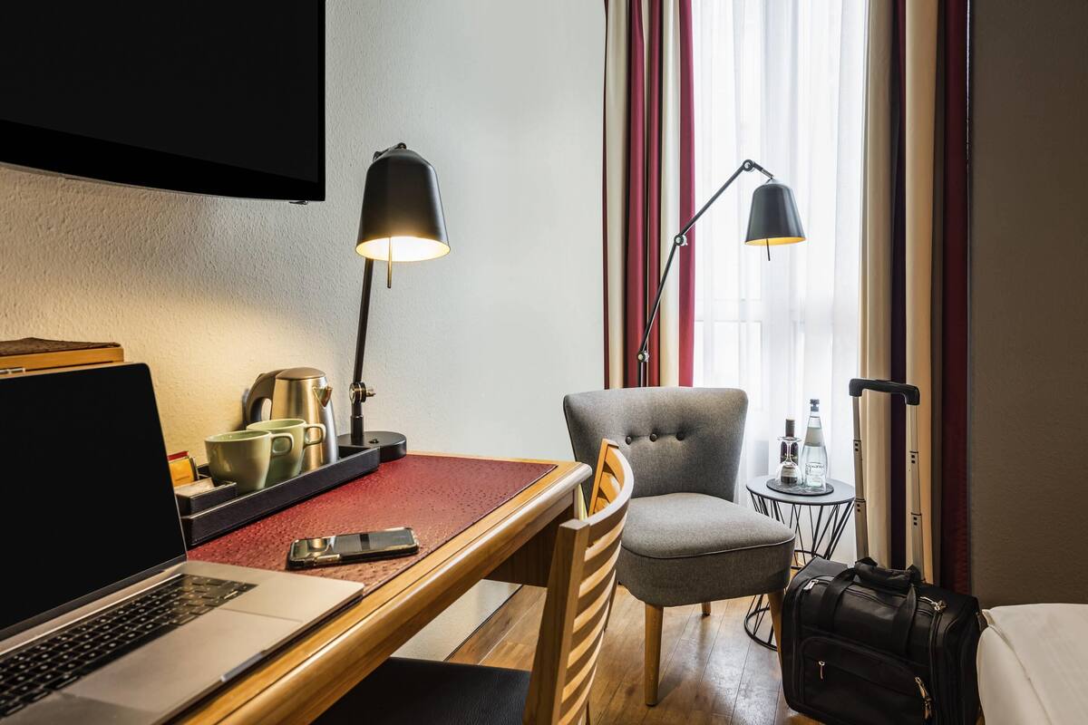 A hotel room with a desk, laptop, chair, bedside table, lamp, and a suitcase.