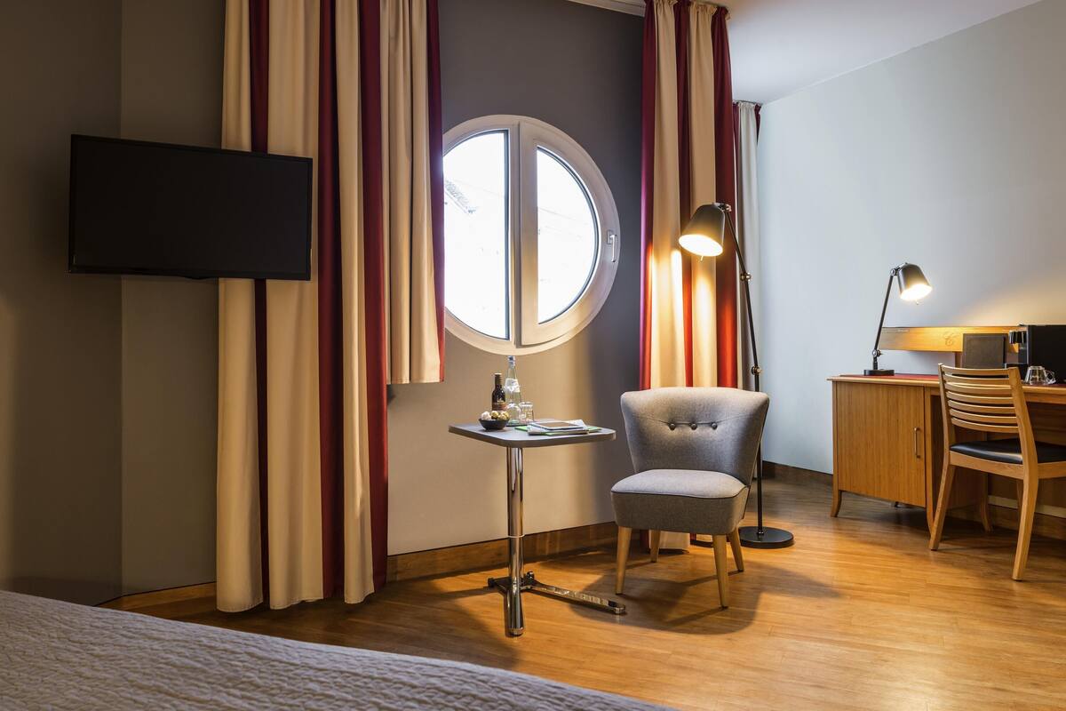 A hotel room with a bed, a desk with a chair, a television mounted on the wall, and a window with curtains.