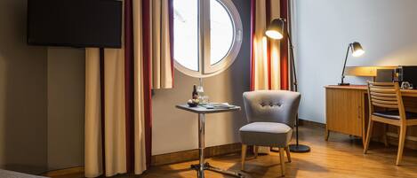A hotel room with a bed, a desk with a chair, a television mounted on the wall, and a window with curtains.