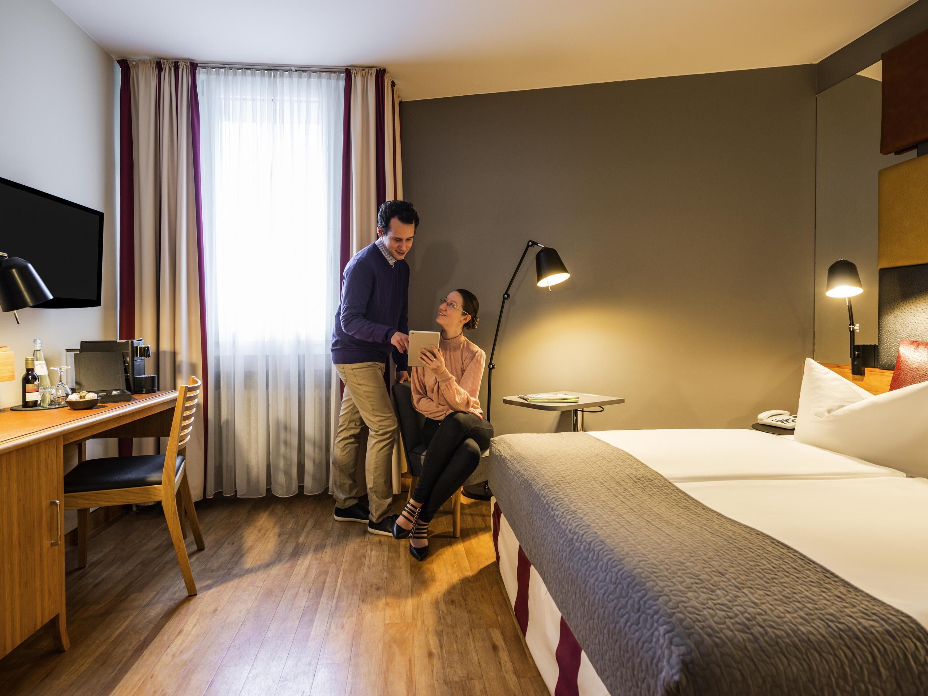 Une chambre d’hôtel équipée d’un lit, d’un bureau, d’une chaise, d’une télévision, d’une lampe et d’une fenêtre avec des rideaux.