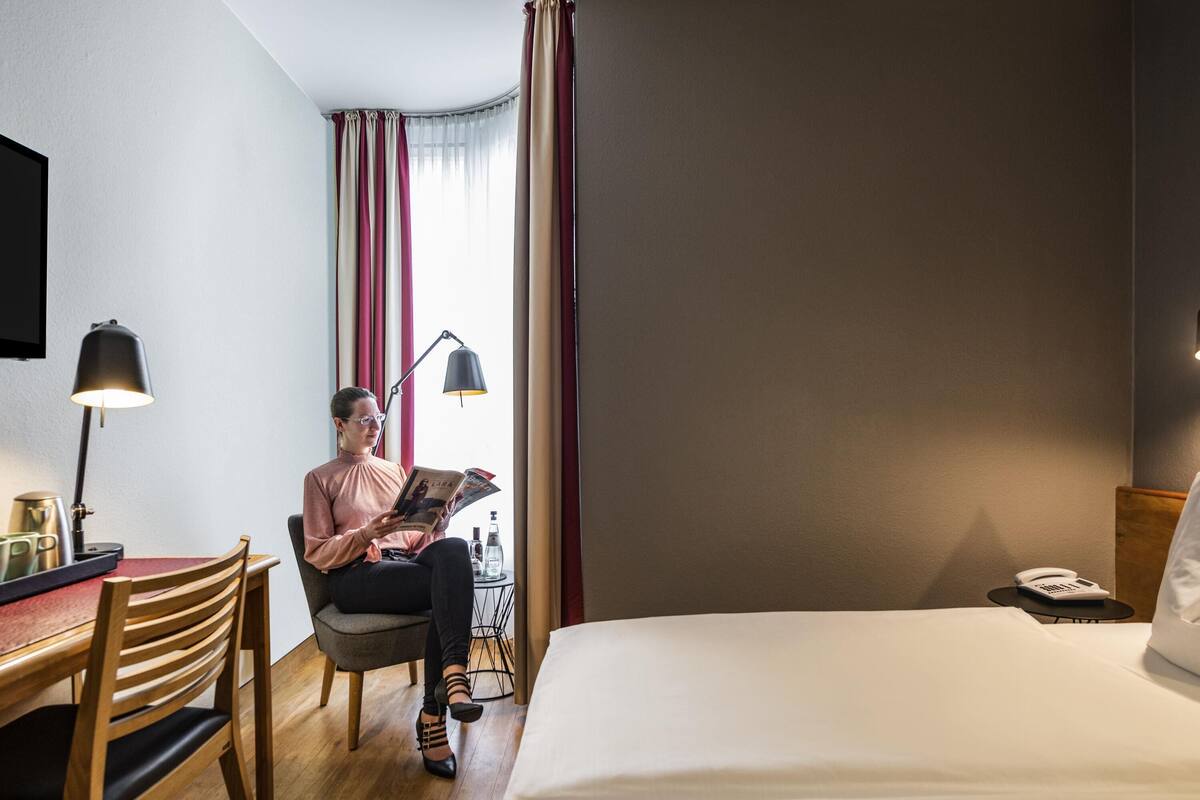 A hotel room with a bed, a desk with a lamp, a chair, and a window with curtains.