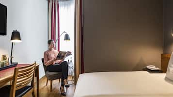 Una habitación de hotel con una cama, un escritorio con lámpara, una silla y una ventana con cortinas.