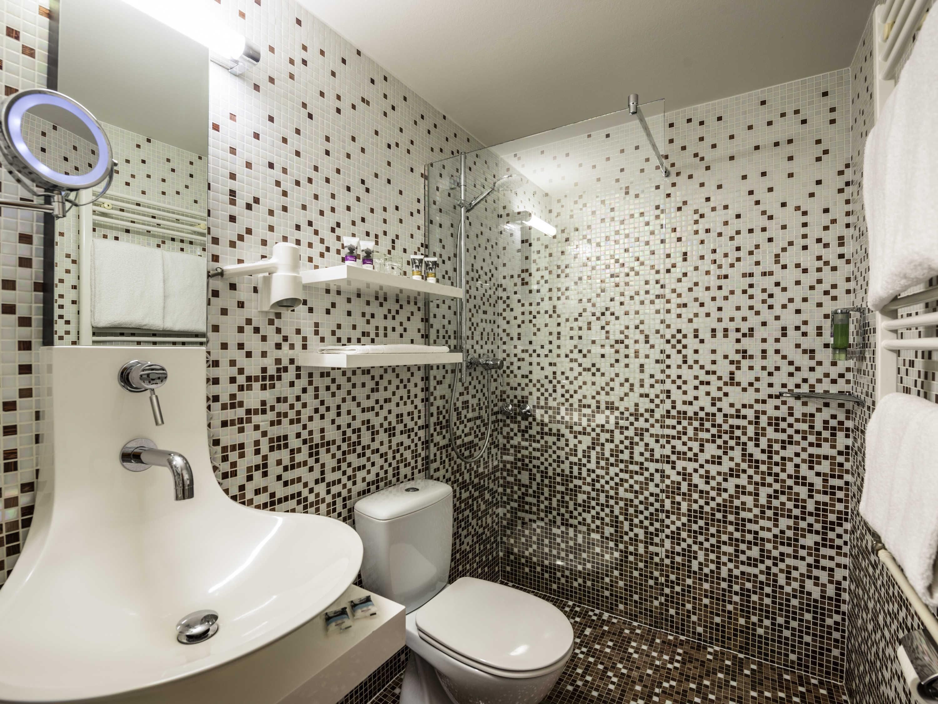 A bathroom with a white sink, toilet, and shower area. There are shelves above the sink and a mirror with a ring light.