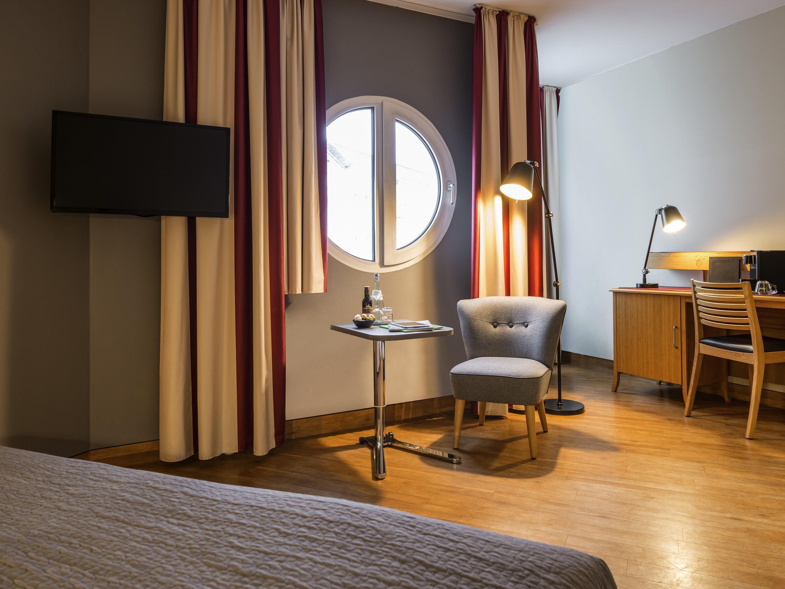 A hotel room with a bed, a desk with a chair, a television mounted on the wall, and a window with curtains.