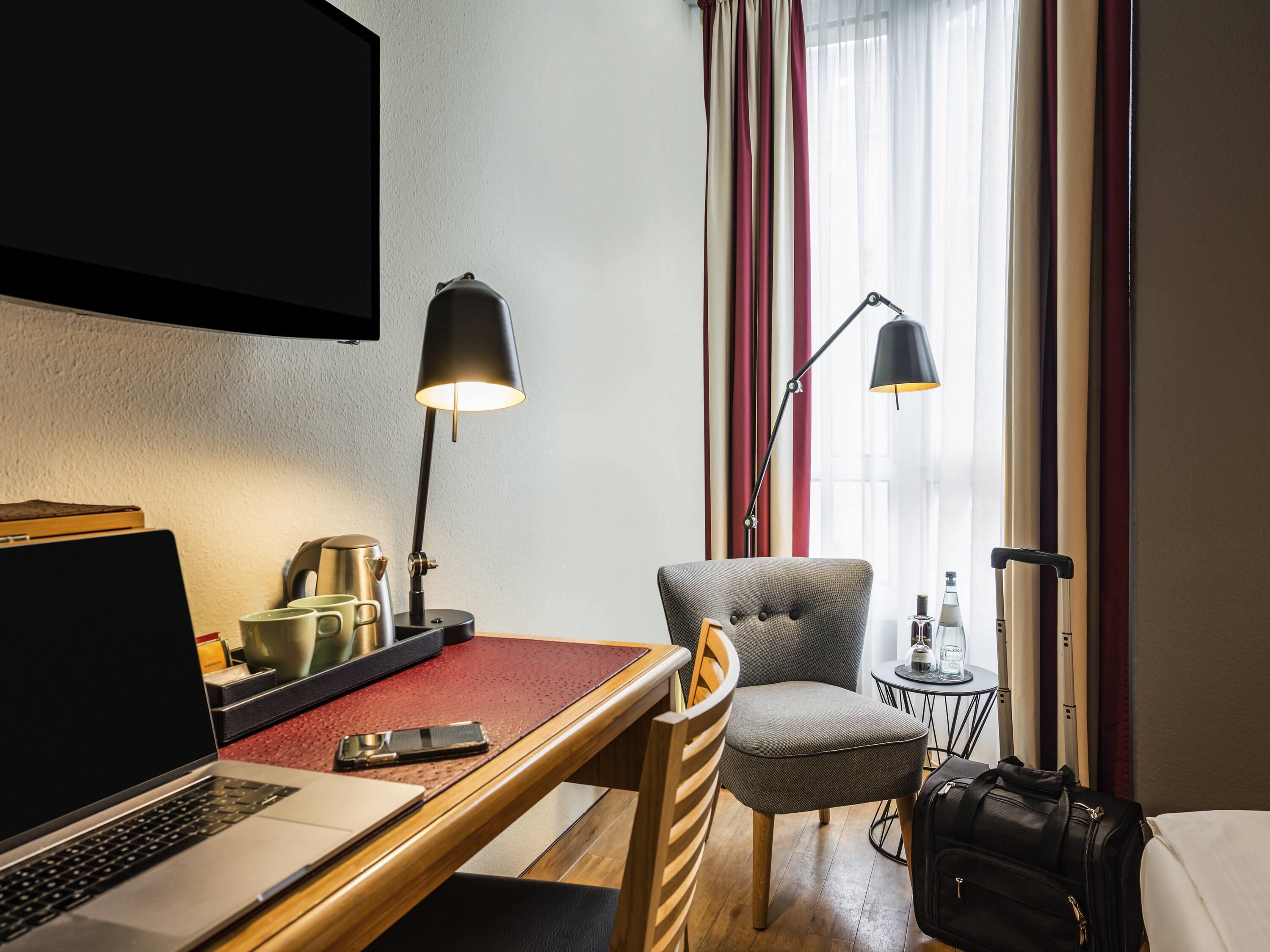 A hotel room with a desk, laptop, chair, lamp, and luggage.