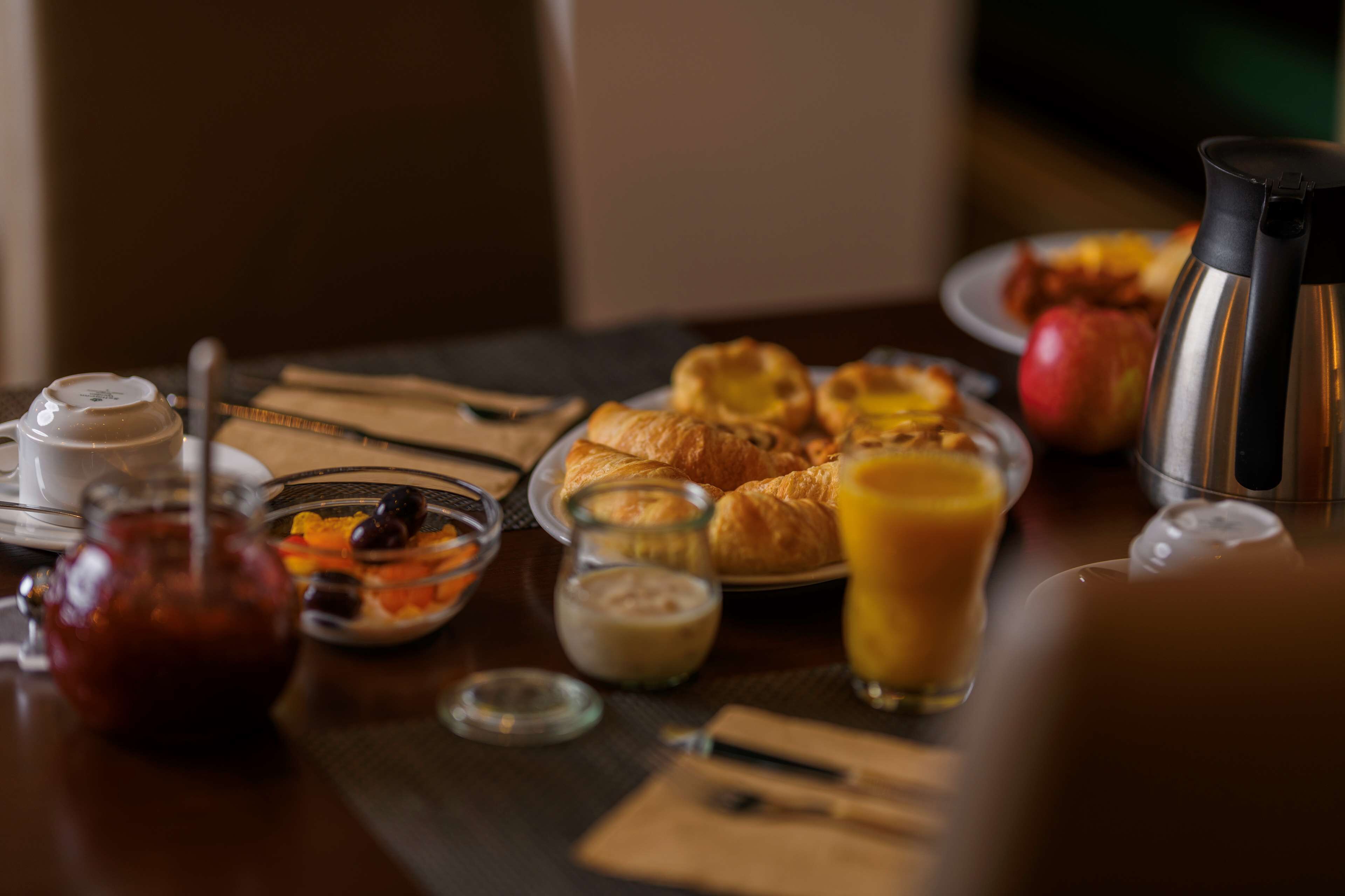 Una mesa de desayuno con cruasanes, un vaso de jugo de naranja, un termo y un bol de fruta.