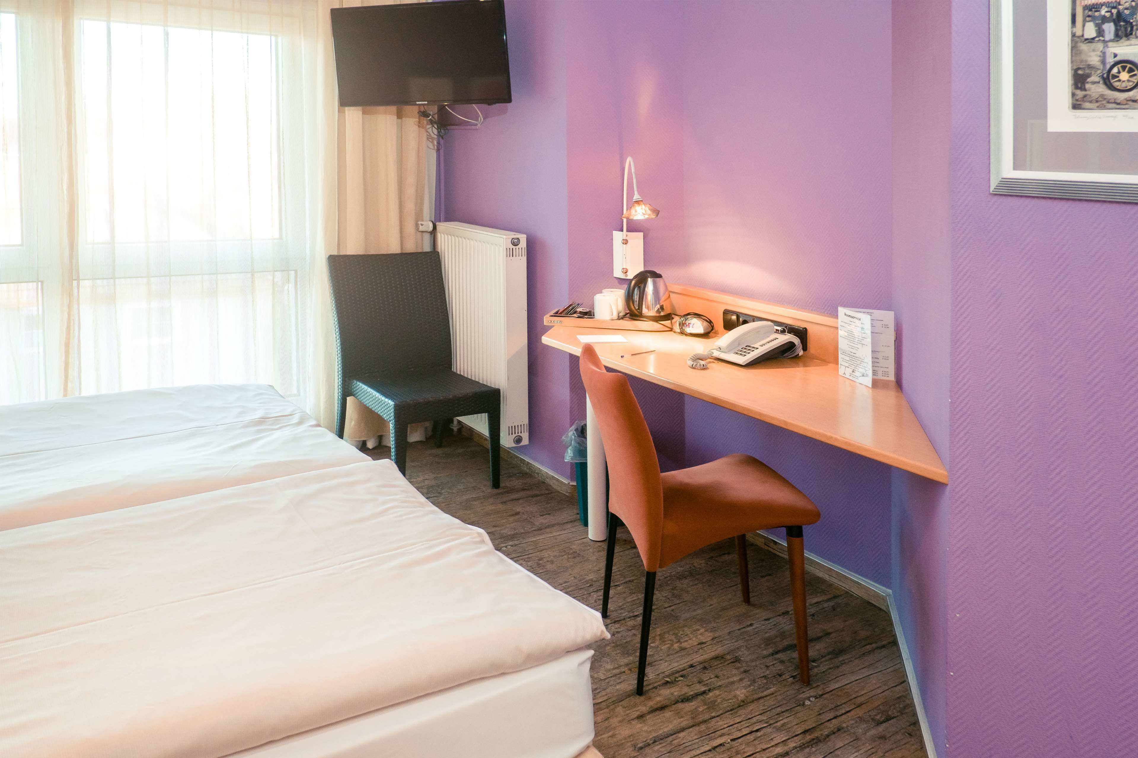 Una habitación de hotel con una cama, un escritorio con silla, televisión y ventana con cortinas translúcidas.