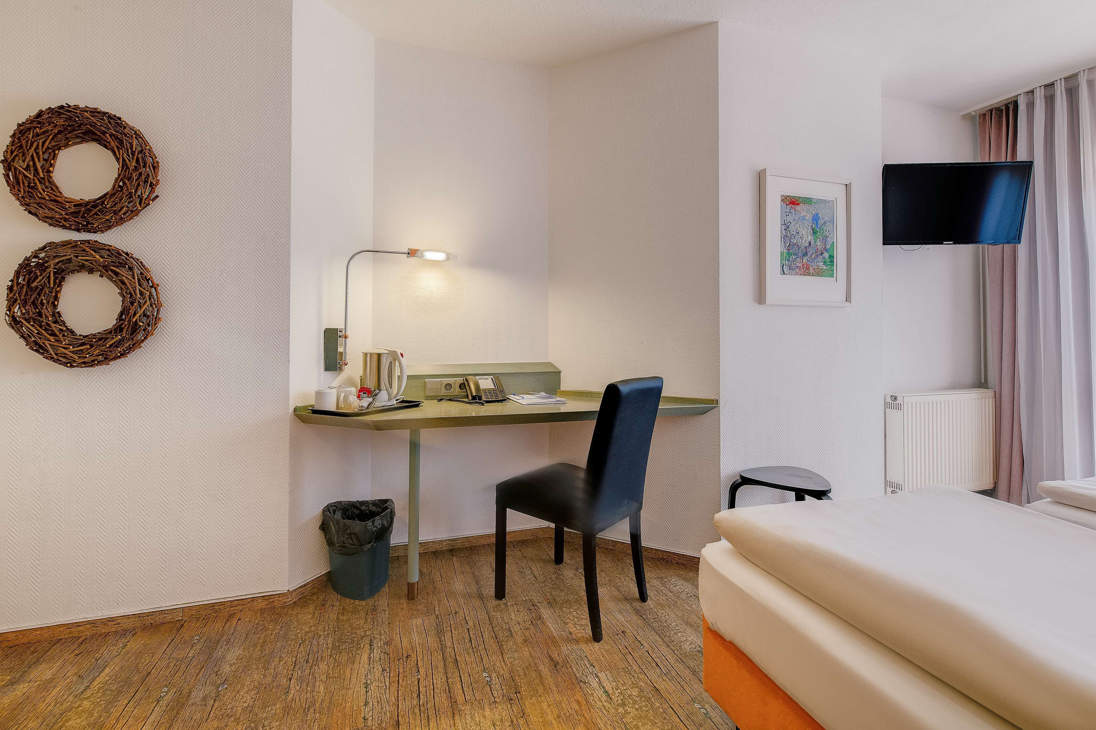 Una habitación de hotel con cama, escritorio con silla, cubo de basura, televisor montado en la pared y dos coronas decorativas.