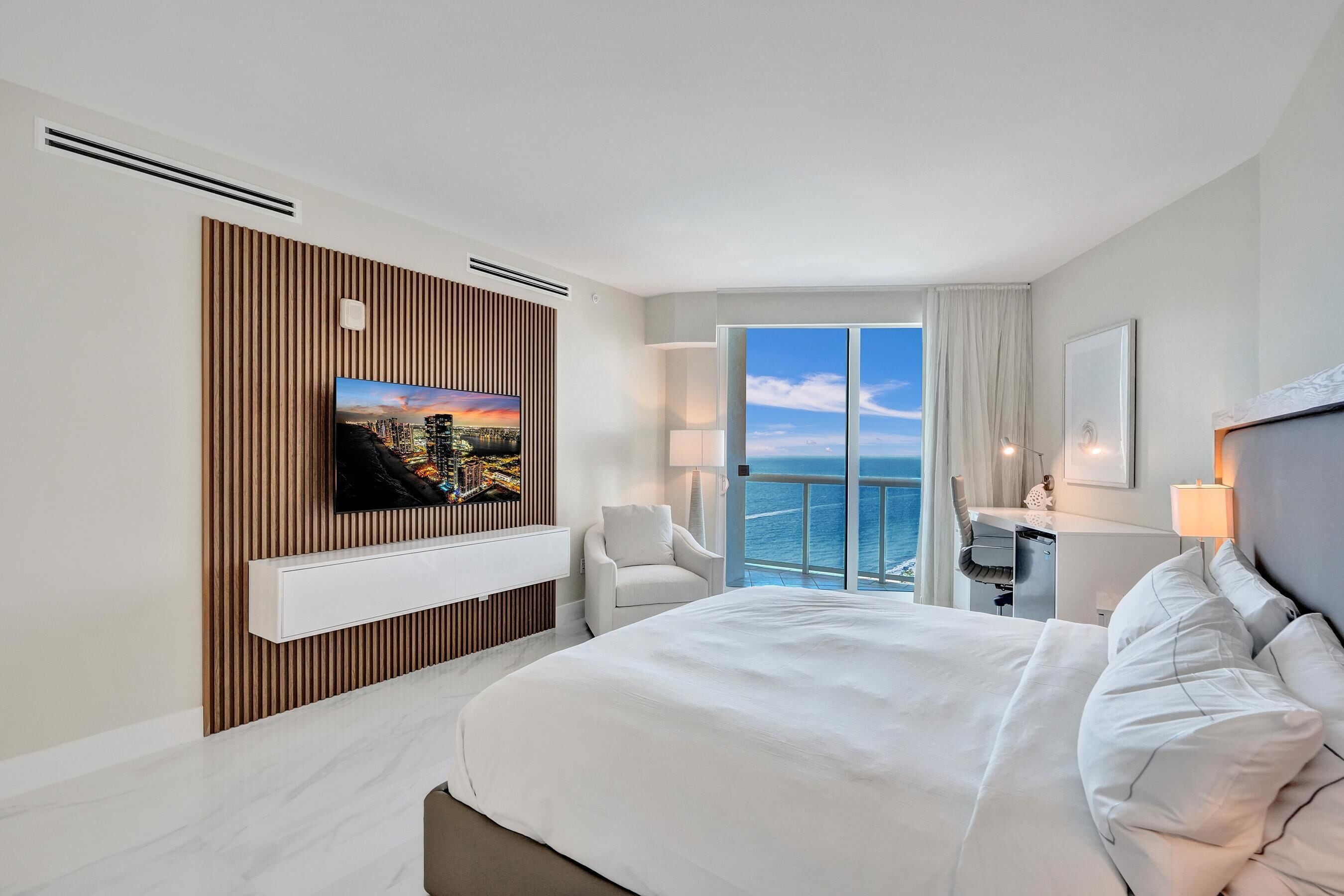 A modern hotel room with a large bed, a TV mounted on a wooden panel wall, a desk with a chair, and a balcony with a view of the sea.
