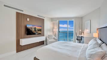 A modern hotel room with a large bed, a TV mounted on a wooden panel wall, a desk with a chair, and a balcony with a view of the sea.