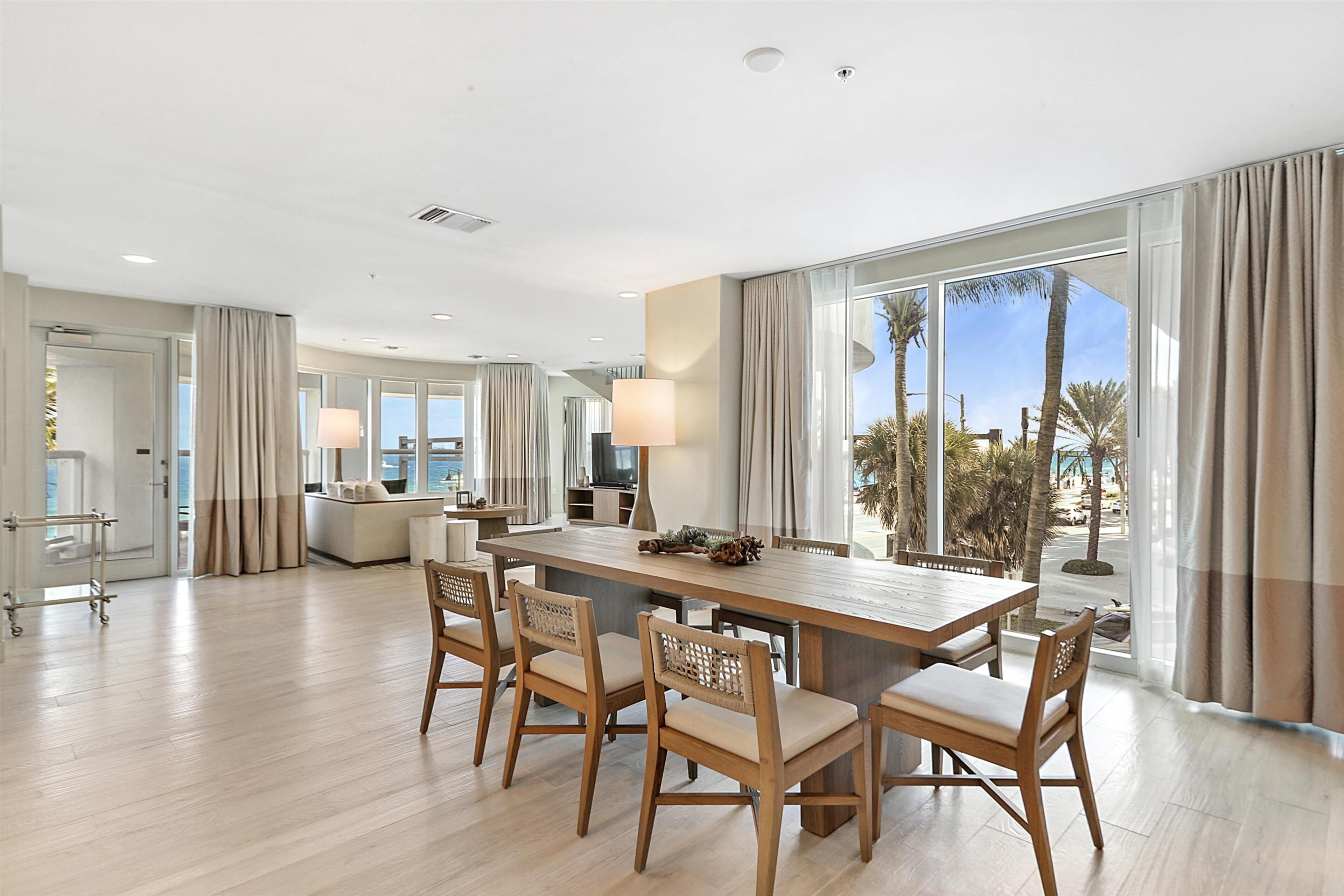 A spacious hotel dining area with a large wooden table and chairs, a modern kitchen in the background, and large windows offering a view of palm trees and the sky.