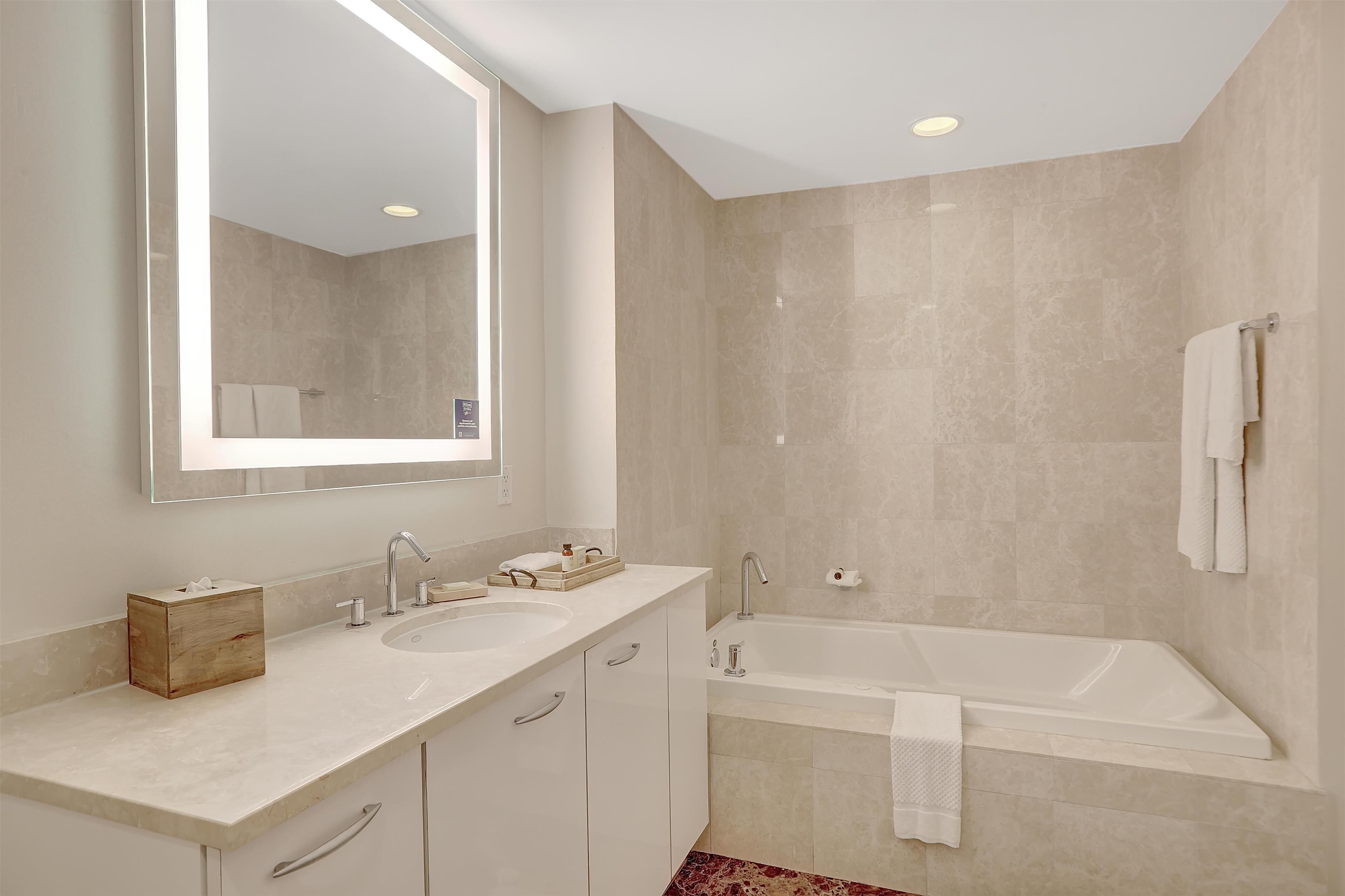 A modern bathroom with two sinks, a large mirror, a bathtub, and beige tiled walls.