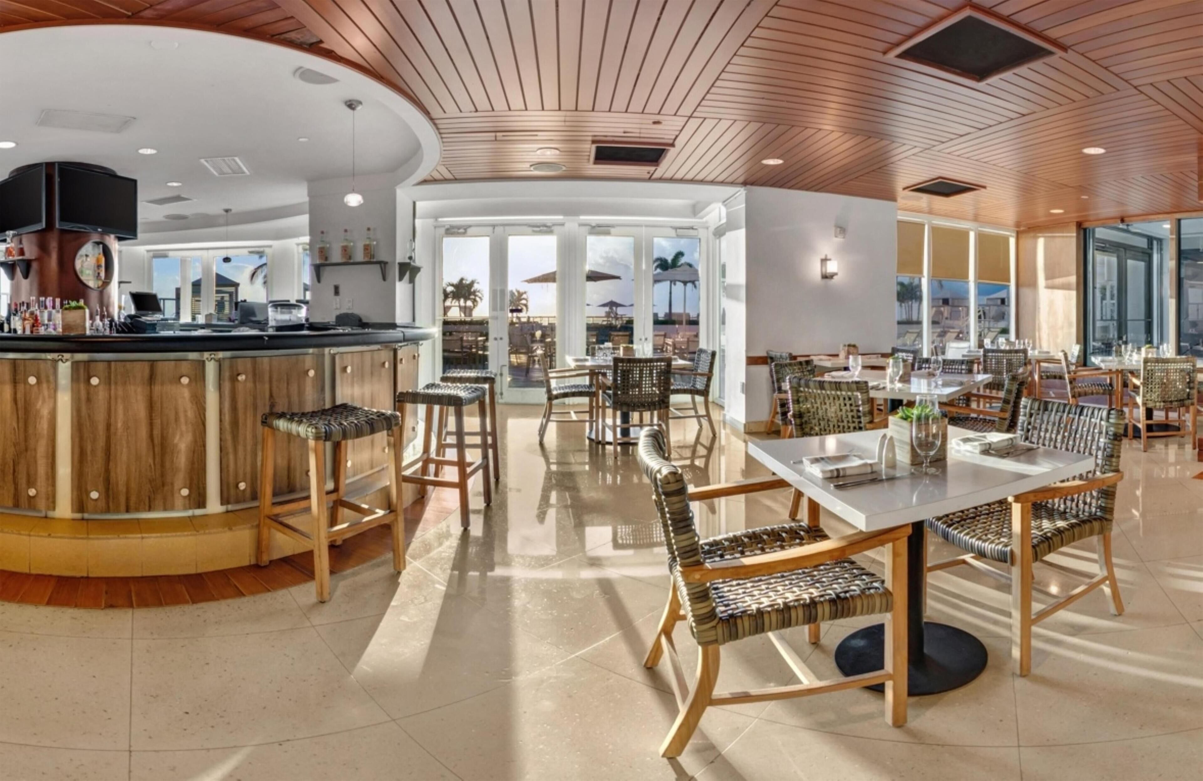 A spacious restaurant with a wooden bar, wicker furniture, and large windows offering views.