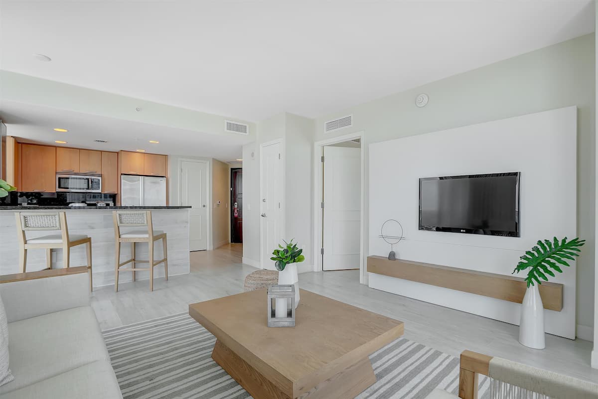 A modern living room with a wooden coffee table, a TV mounted on the wall, and a kitchen in the background.