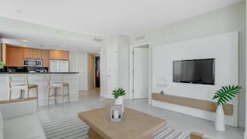 A modern living room with a wooden coffee table, a TV mounted on the wall, and a kitchen in the background.