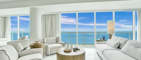 A modern living room with a large round wooden coffee table, two white sofas, and a view of the ocean through large windows.