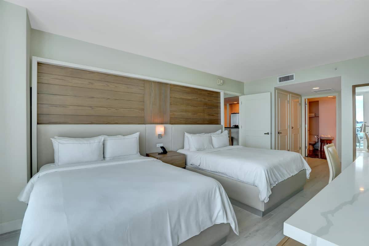 A hotel room with two beds, a wooden headboard, a bedside table with a lamp, and a view of the bathroom and dining area.