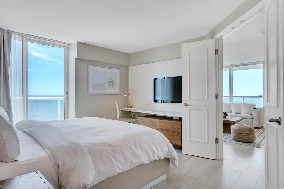 A modern bedroom with a bed, a desk, a TV, and a view of the sea.