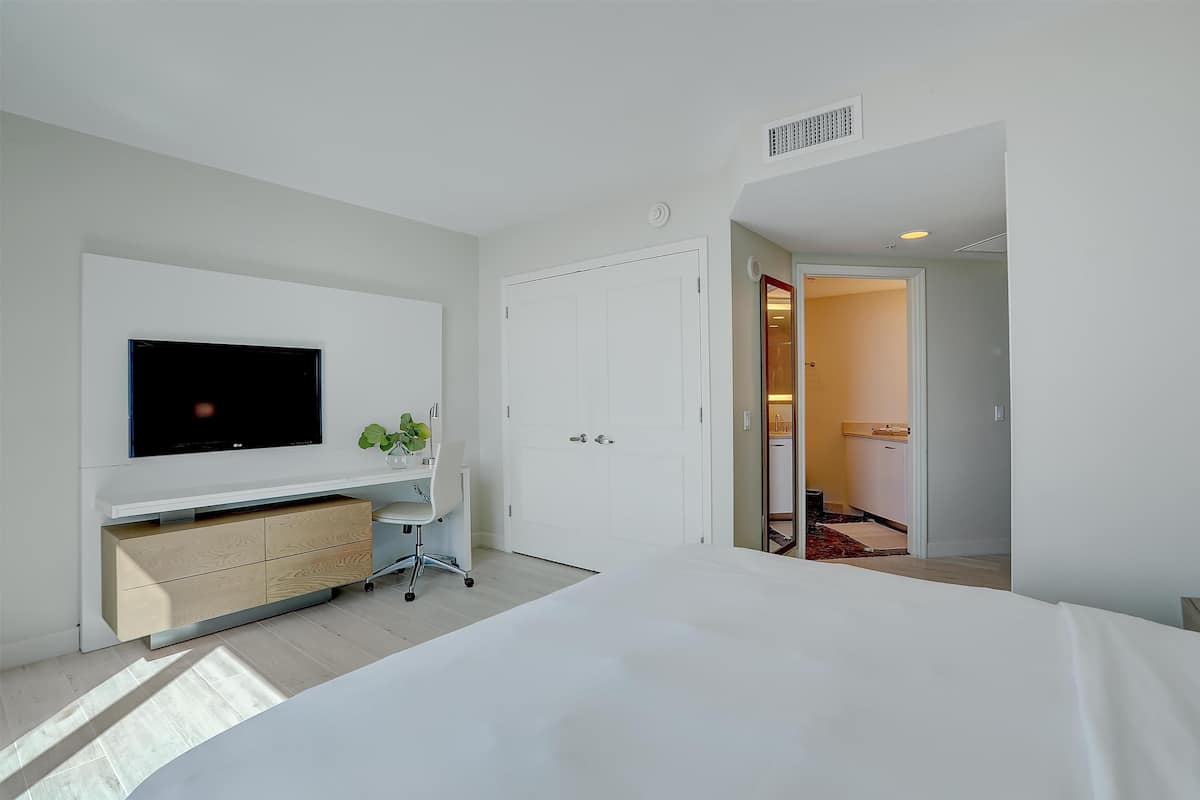 A modern hotel room with a large bed, a TV mounted on the wall, a desk with drawers, and a door leading to a bathroom.