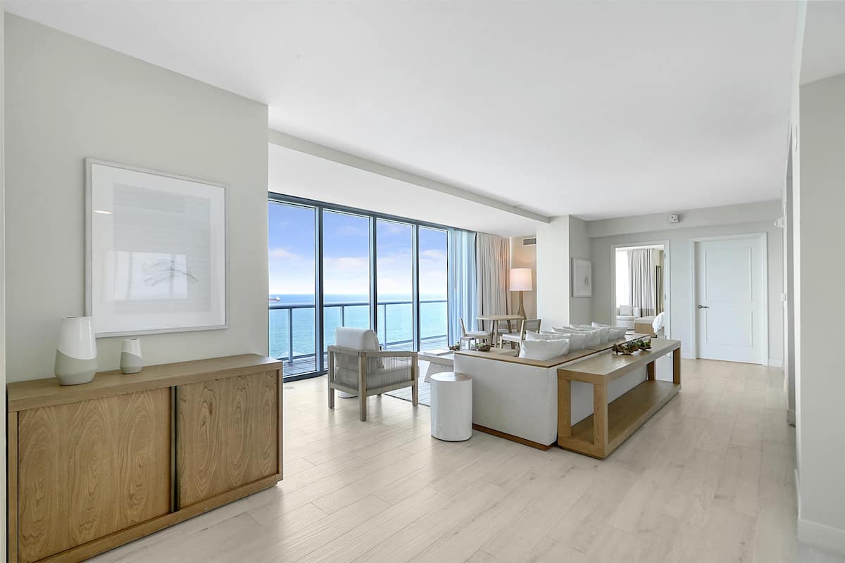 A modern hotel room with a large window, a wooden cabinet, a seating area, and a central table with a mirror.