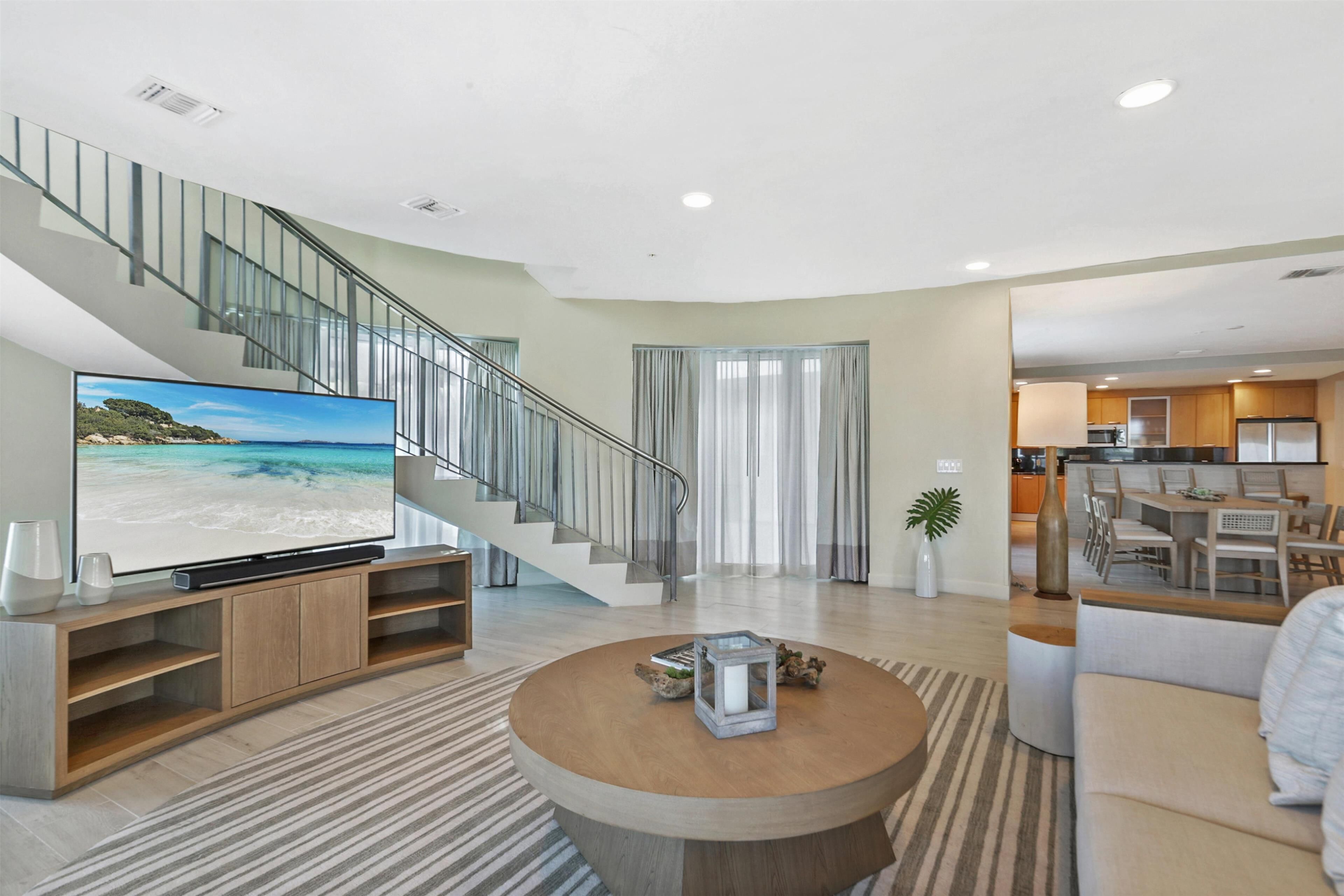 Una sala moderna con un televisor de pantalla plana, una mesa de centro redonda, una escalera y una cocina visible a través de una puerta abierta.
