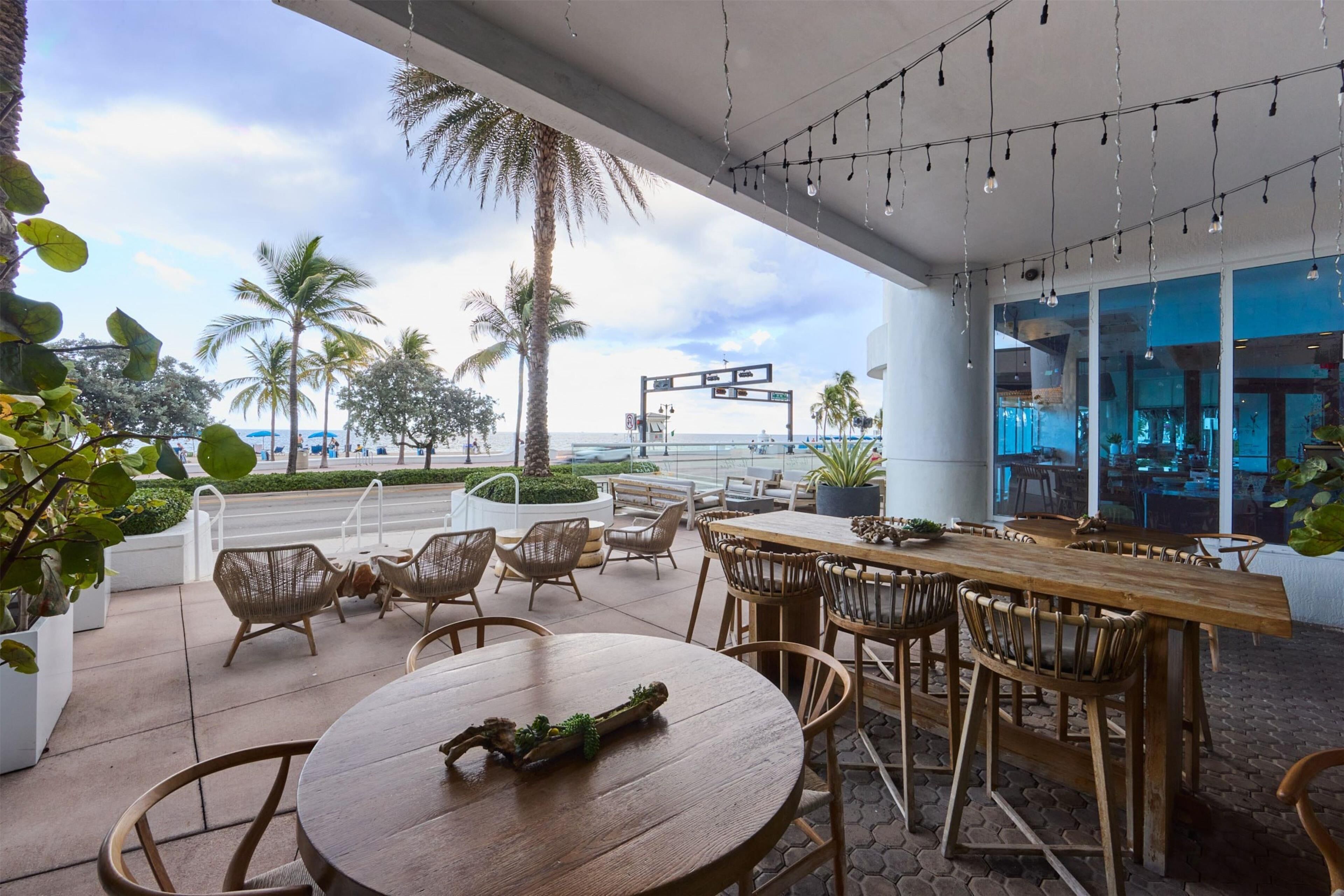 Un espace extérieur aménagé avec des tables et des chaises en bois, offrant une vue sur une plage et des palmiers.