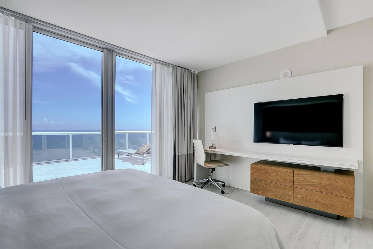 A modern hotel room with a large bed, a desk with a chair, a TV mounted on the wall, and a balcony with a view of the sky.