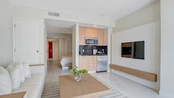 Uma sala de estar moderna com sofá, mesa de centro com um vaso, área de cozinha com micro-ondas e geladeira, e uma TV fixada na parede.