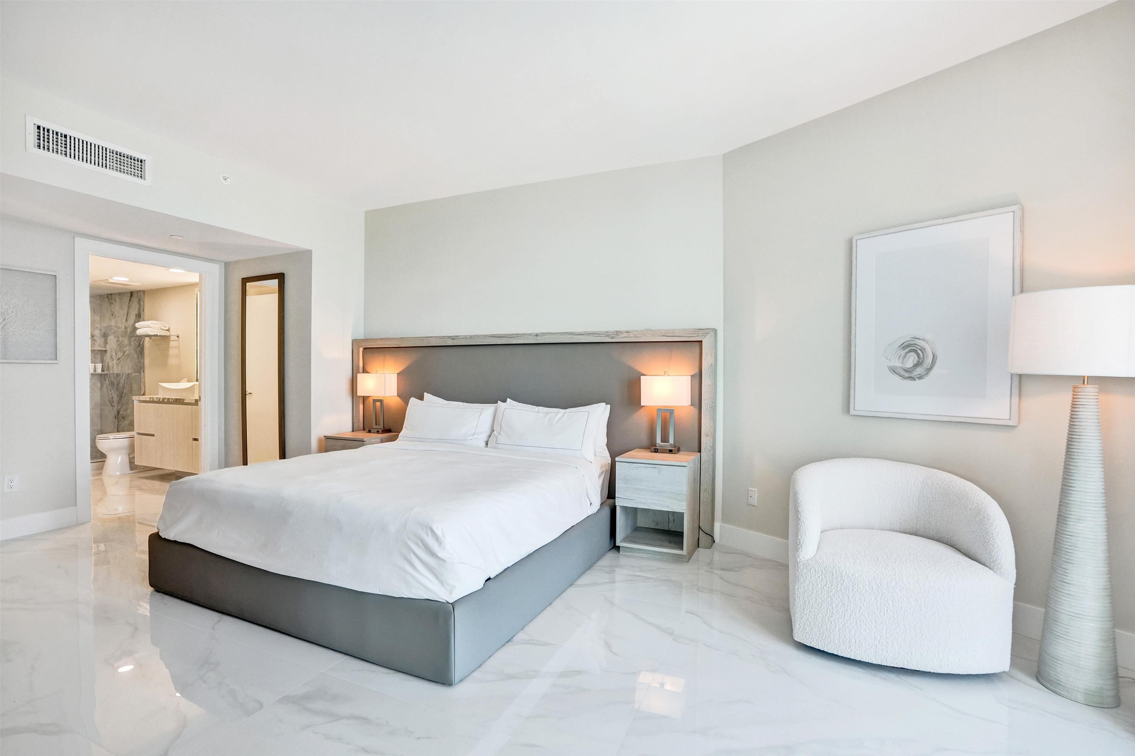A modern hotel room with a large bed, bedside lamps, a white armchair, and a framed picture on the wall.
