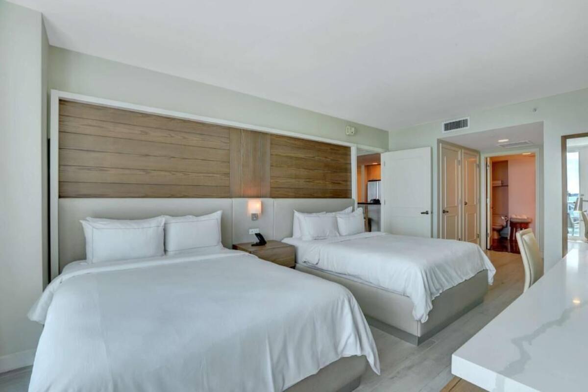 A hotel room with two beds, a wooden headboard, a bedside table with a lamp, and a view of the city.