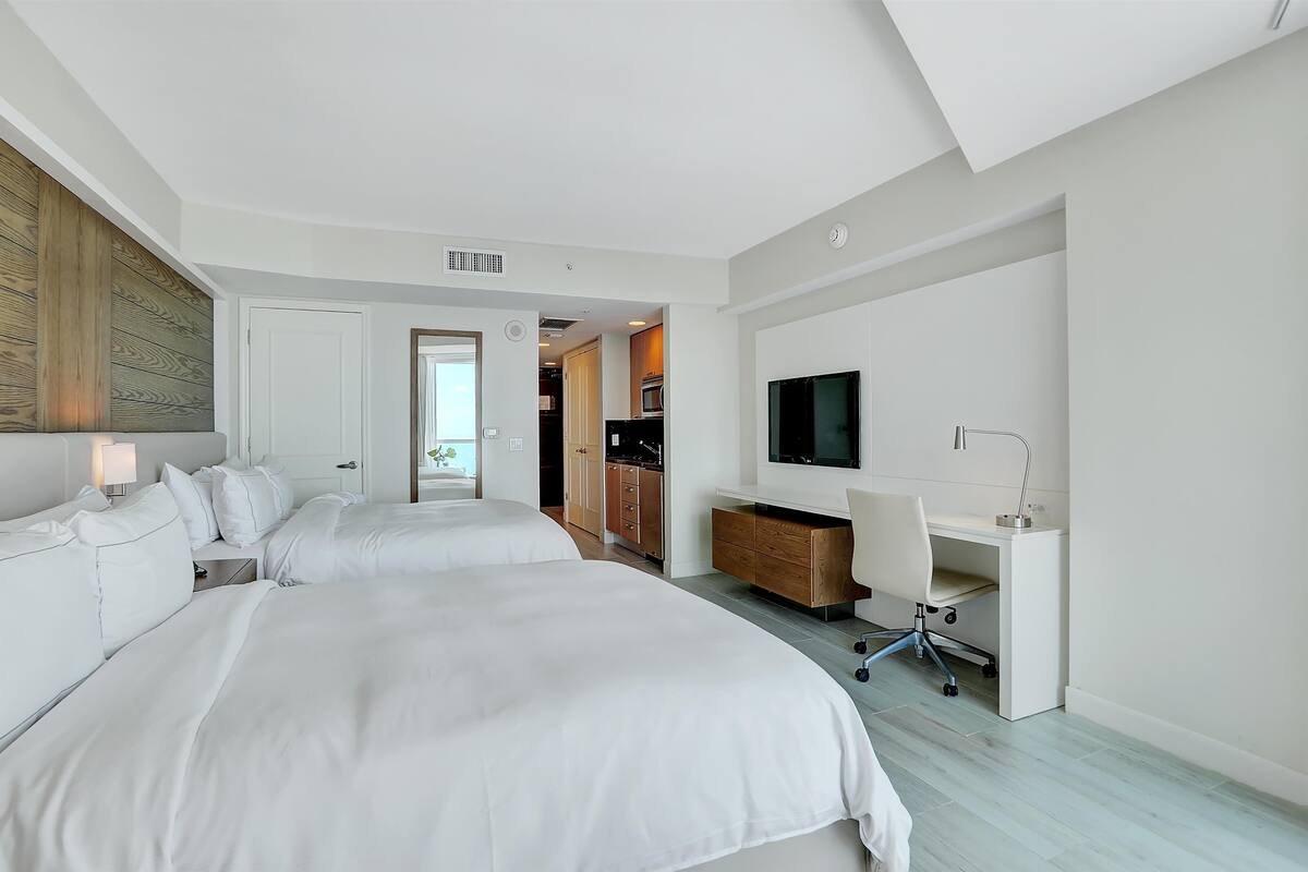 A modern hotel room with two beds, a desk, a TV, and a kitchenette.