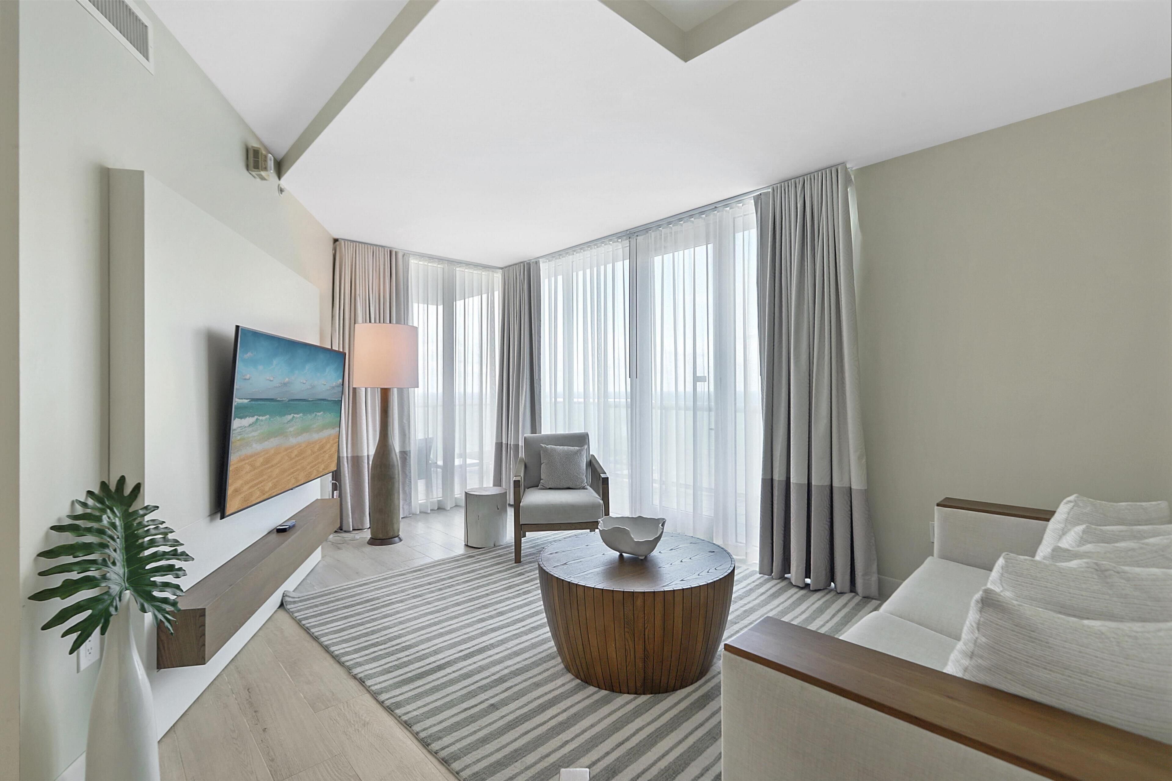 A modern hotel room with a large window, a flat-screen TV, a comfortable sofa, a round coffee table, and a side table.