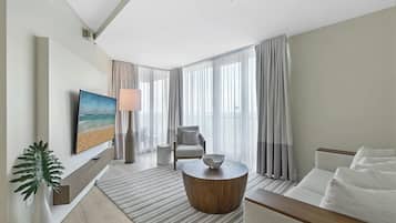 A modern hotel room with a large window, a flat-screen TV, a comfortable sofa, a round coffee table, and a side table.