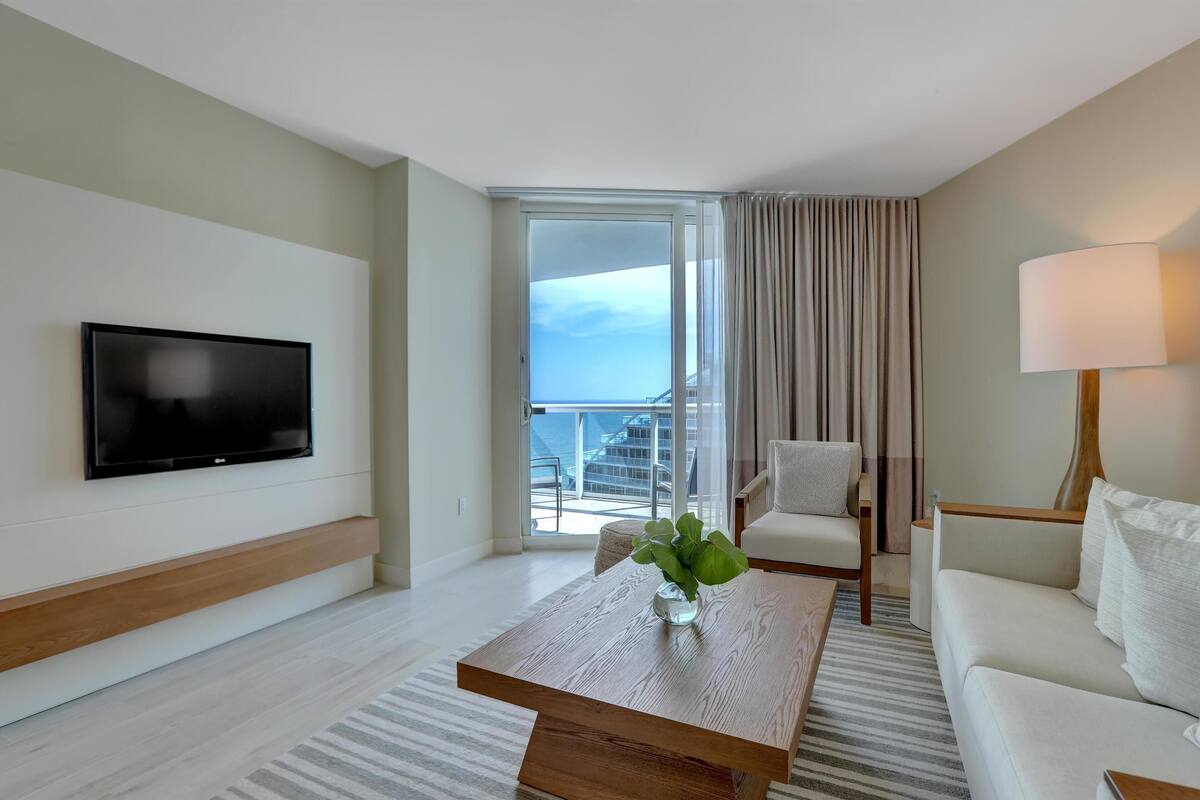 A modern hotel room with a flat-screen TV, a wooden coffee table with a vase, a sofa, a chair, and a large window with a view of the sea.