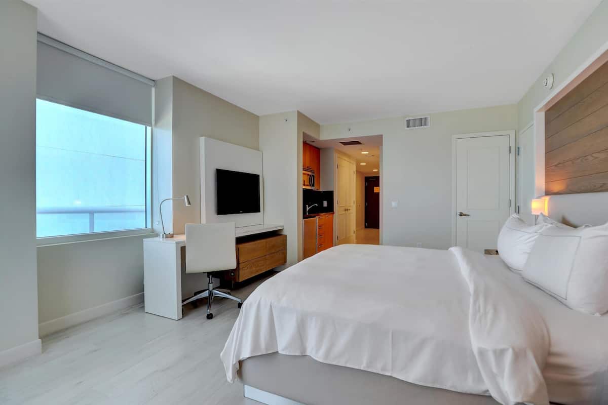 A modern hotel room with a large bed, a desk with a computer, a TV, and a window providing a view of the city.