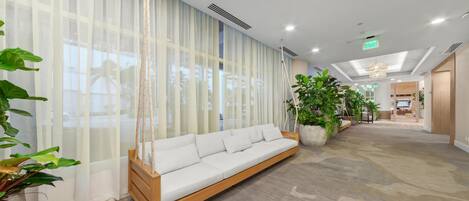 A modern lobby with a white sofa, large windows with sheer curtains, and potted plants.