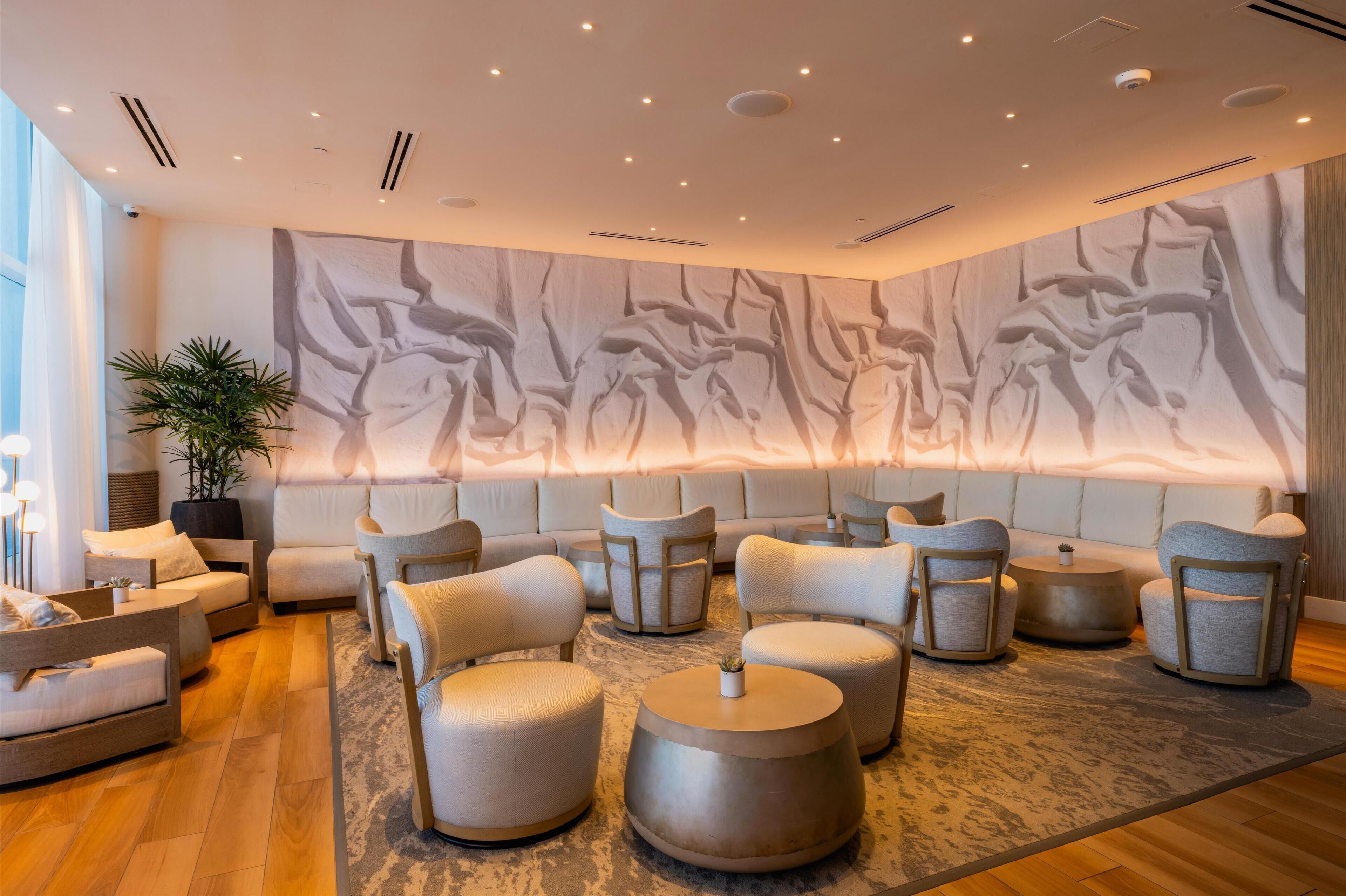 A modern lounge area with sofas, armchairs, and round tables, featuring a large abstract wall mural and a potted plant.