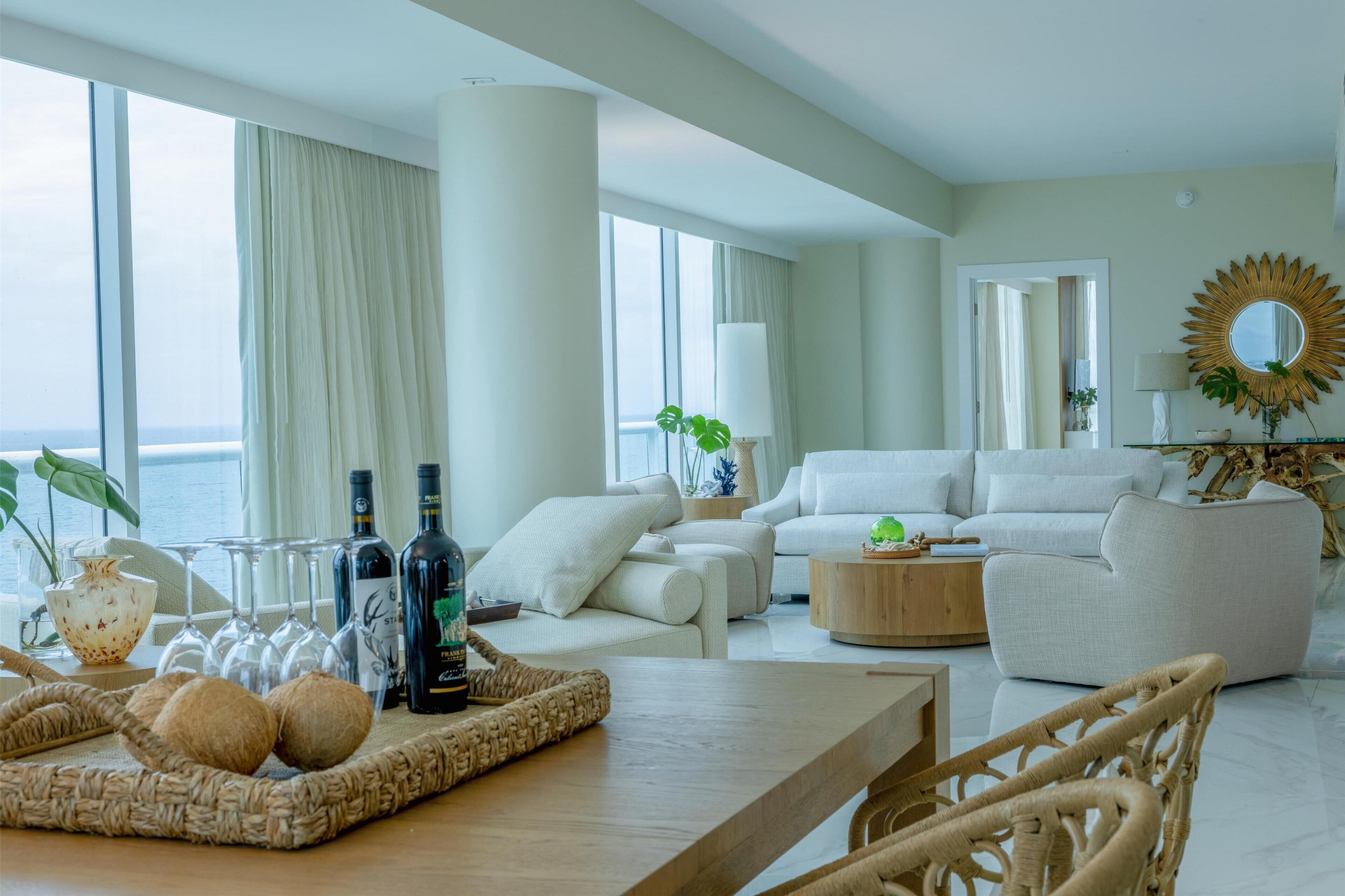 A modern living room with a large window, white sofas, a wooden coffee table, and a wicker chair.