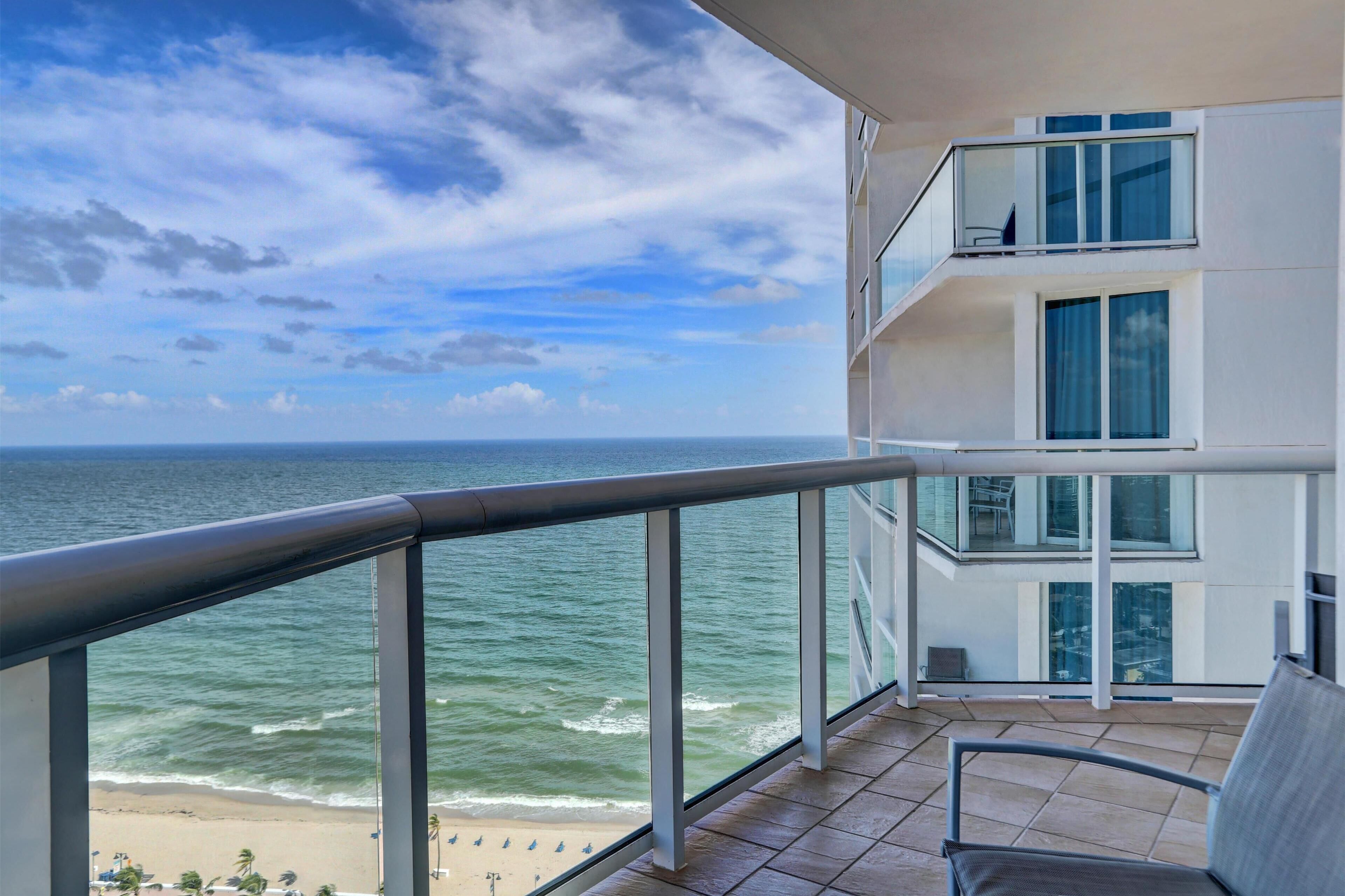 A balcony with a view of the ocean and beach, featuring a modern building with multiple floors and large windows.