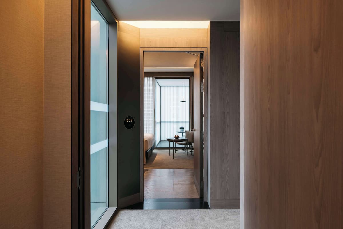A hotel hallway leading to a room with a bed, a small table, and a chair.