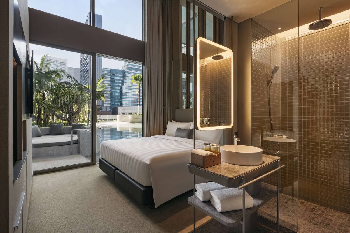 A modern hotel room with a large bed, a bathroom with a glass-enclosed shower, and a view of palm trees and buildings.