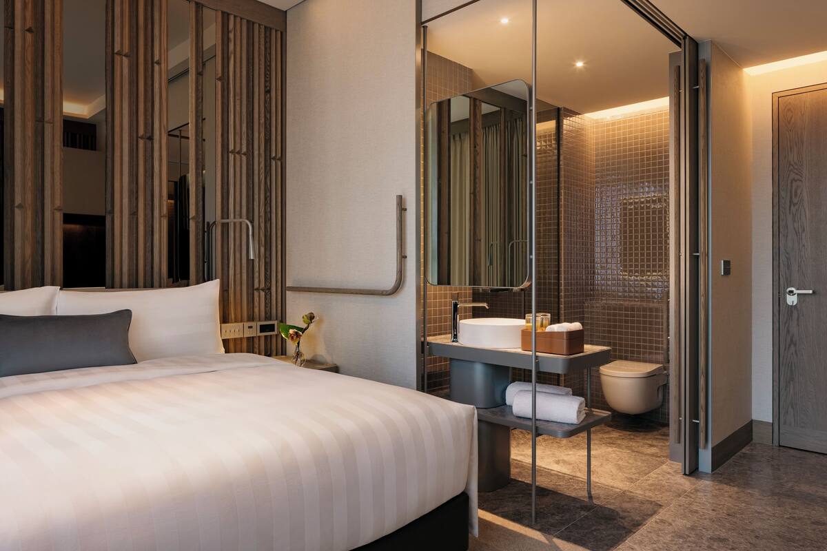 A modern hotel room with a large bed, a bathroom with a glass partition, and a wooden panel wall.
